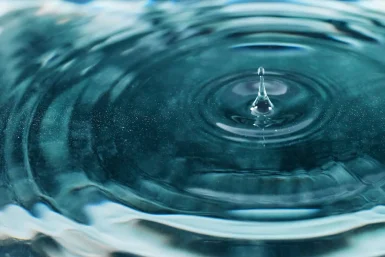 Close-up of a transparent blue water droplet creating ripples on the surface, capturing the moment of impact in soft light.